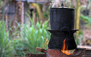 Black pot boiling for the rice cooker on the fired stove next to firewood pile.