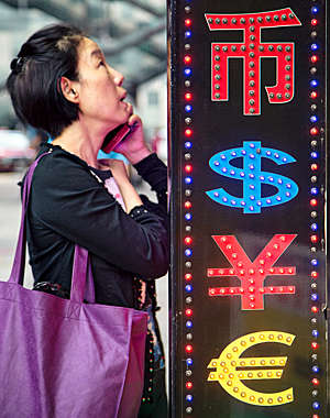 Asian woman talks on her smartphone next to a neon sign displaying currency codes. 