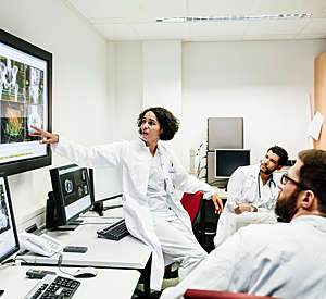 Female clinical registrar perched sideways on a desk discusses with two male colleagues a patient's test results displayed on a large screen.