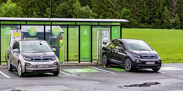 Two cars plugged into an electric vehicle charging station against the green backdrop of the Norwegian countryside.