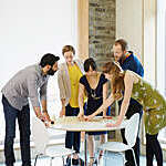 Five colleagues pore over a workplan displayed on a small round table during an animated brainstorming session.