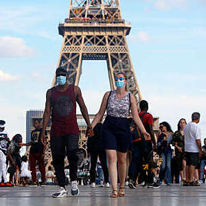 Tourist couple wearing protective masks walk along the Eiffel Tower esplanade in Paris holding hands.