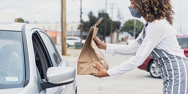 Café attendant, masked, holds out a paper bag of food to a driver in a car.
