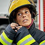 Head and shoulders close-up of late 30s Hispanic first responder with sooty face wearing turnout gear and securing helmet chin strap.