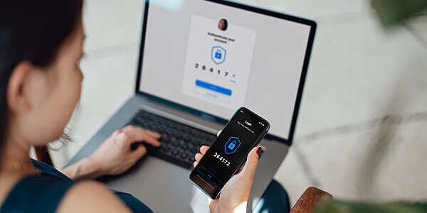 Over the shoulder view of young Asian woman using laptop, logging in online banking account with digital security mobile device at office. Internet security and digital privacy protection concept. Two-Factor Authentication (2FA) concept.